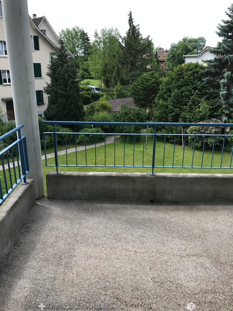 Balcony with gravel flooring, blue metal railing, and a view of a green lawn and surrounding trees.