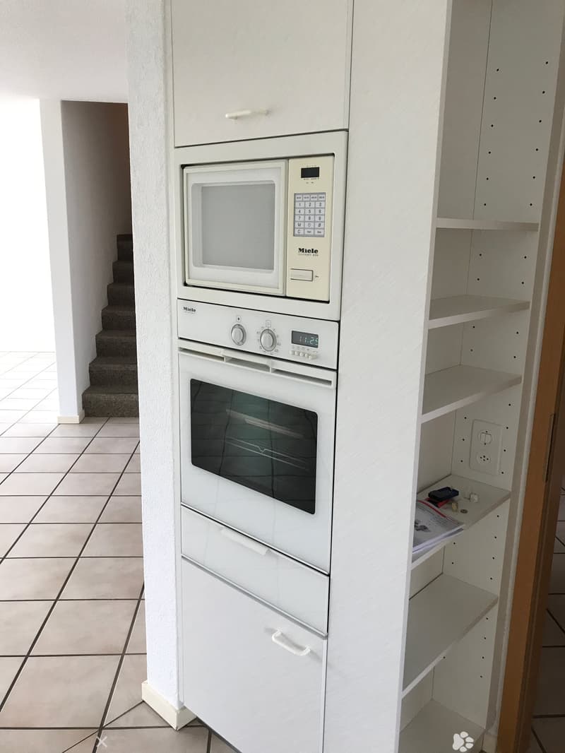 Modern kitchen with white cabinets, a microwave, and an oven. The floor is tiled, and there is a small shelf with some items on it.