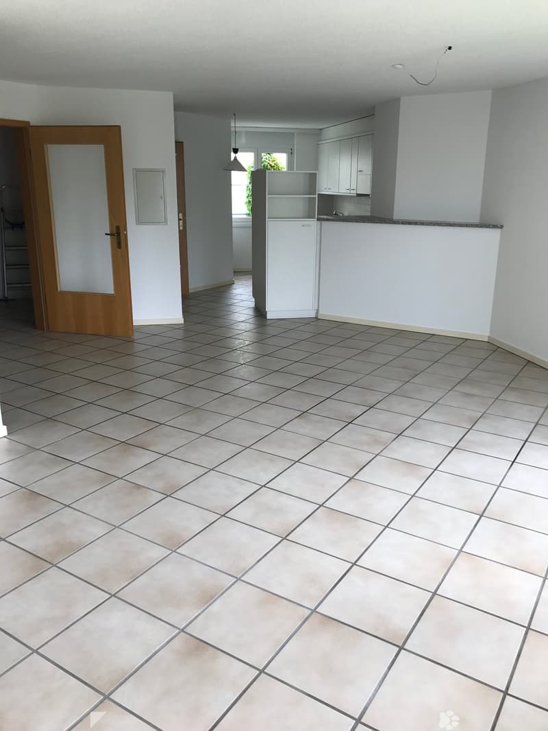 Empty room with tiled floor, white walls, and a doorway leading to another room. The adjacent area shows a small kitchen with white cabinets and a countertop.