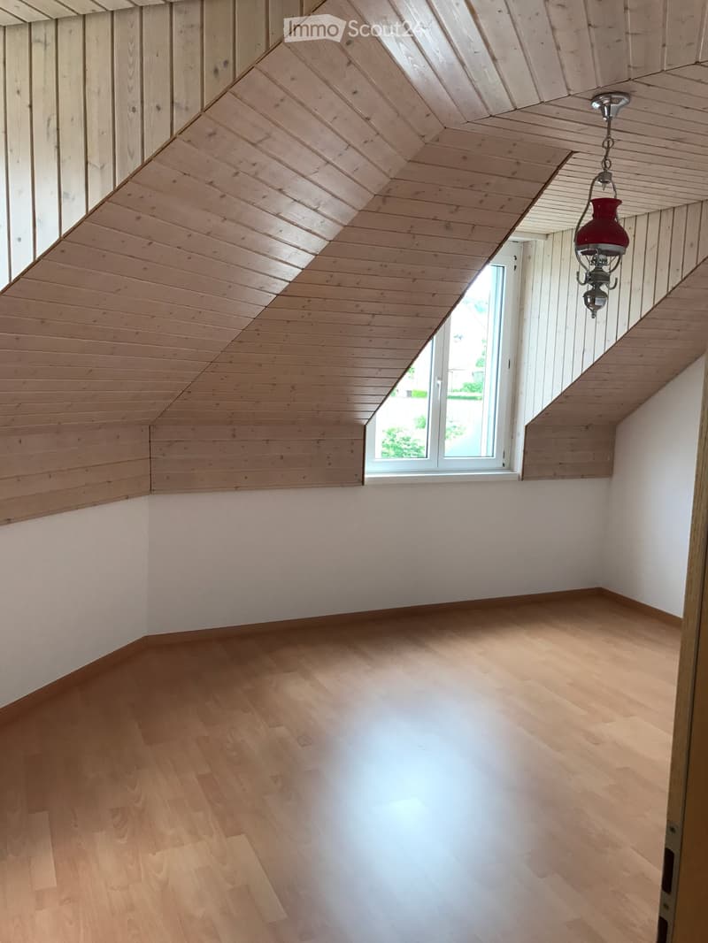 Empty room with wooden floor and slanted wooden ceiling, featuring a single window and a hanging red lantern light fixture.