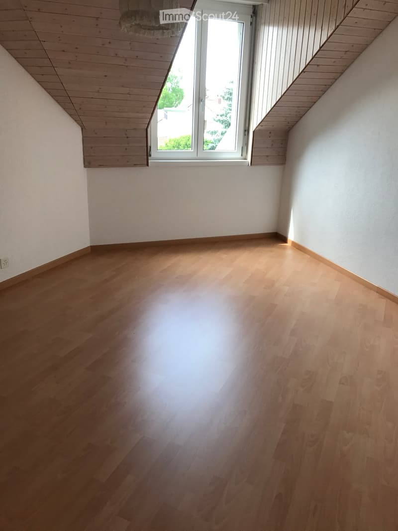 Empty room with wooden flooring and white walls, featuring a large window with white frame and a slanted wooden ceiling.