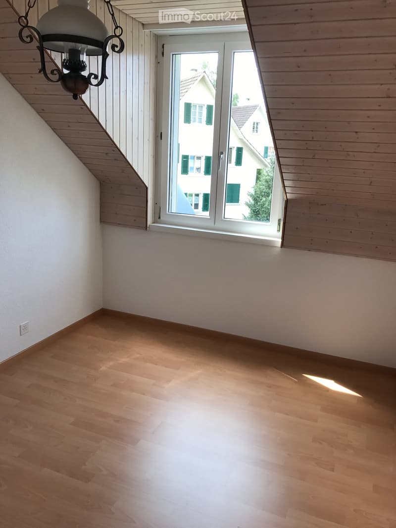 Empty room with wooden flooring, white walls, and a large window offering a view of a neighboring house.
