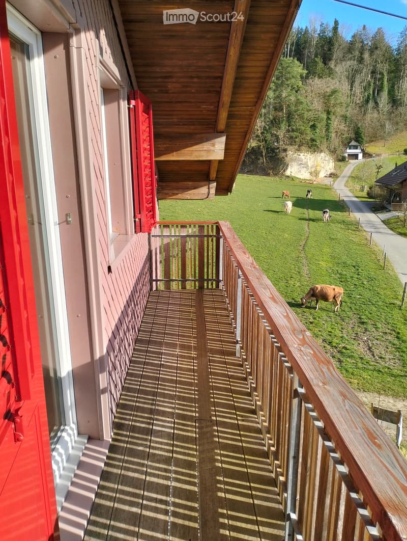 Wooden balcony with railing overlooking a green field with grazing cows and a forested area in the background.