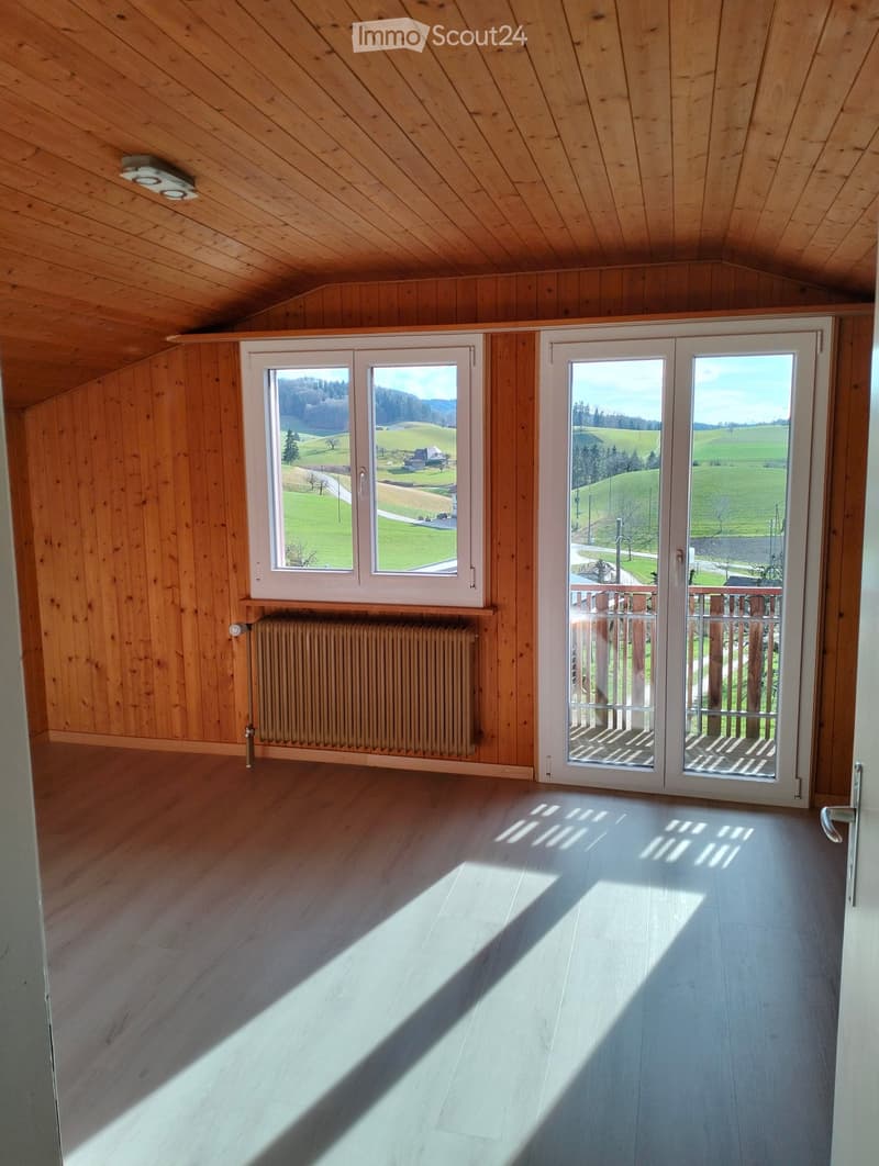 Empty room with wooden walls and ceiling, large windows, and a radiator. Sunlight streams in from the windows, casting shadows on the floor.