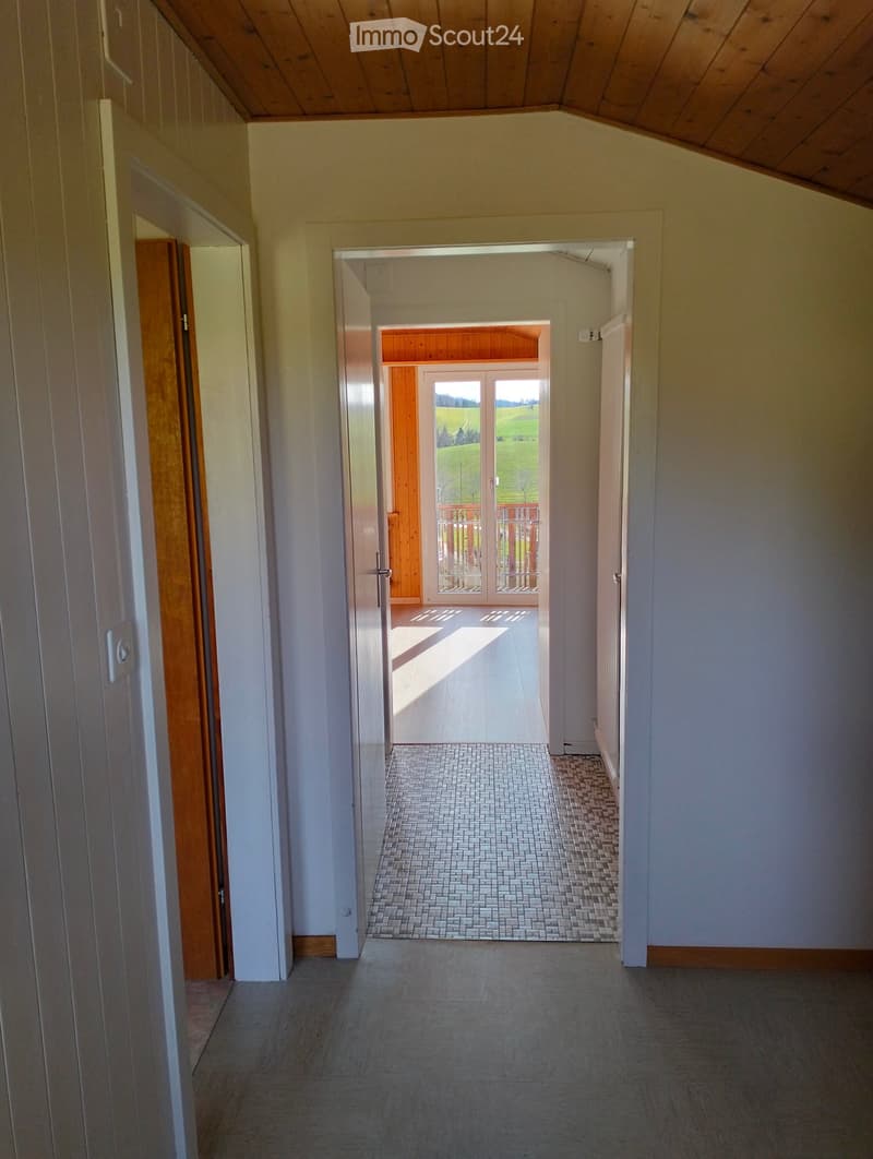 Empty hallway with tiled floor, white walls, and wooden doors leading to a room with a view of a green landscape.