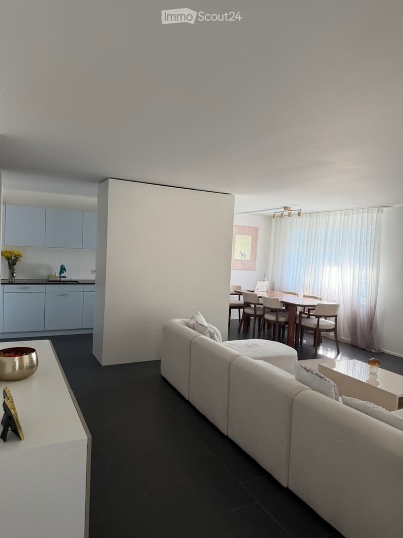 Modern open-plan living and dining area with a white sofa, coffee table, ottoman, dining table with chairs, and a small kitchen area with cabinets and a sink.
