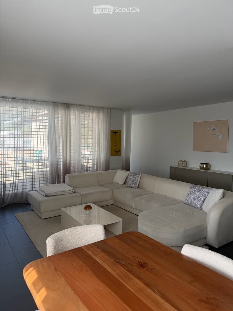 Modern living room with a large sectional sofa, coffee table, wooden dining table, and sideboard. Large windows with sheer curtains allow natural light to fill the space.