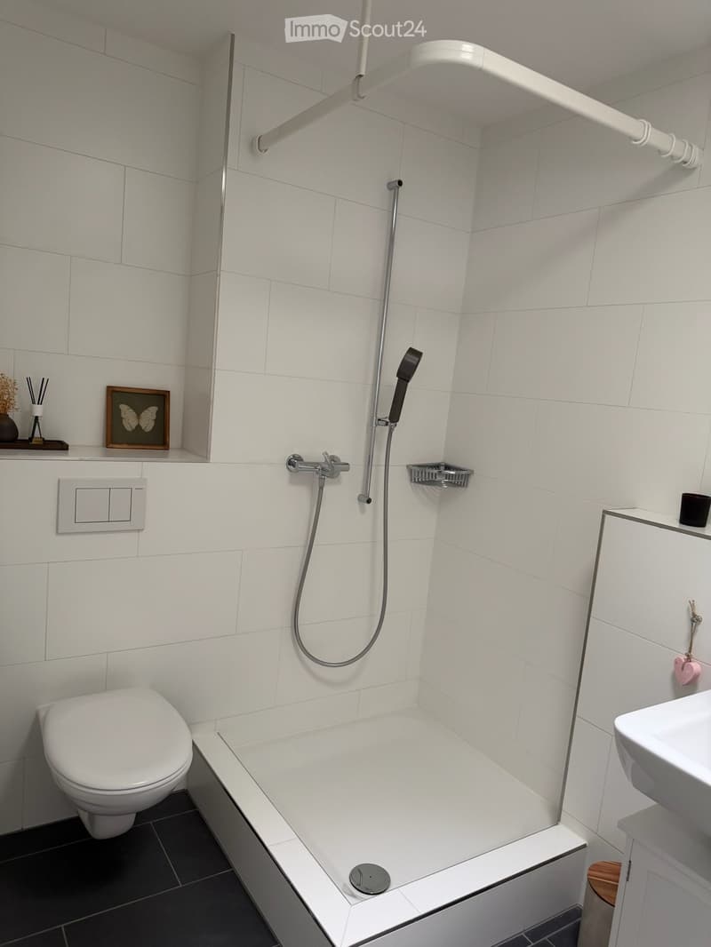 Modern bathroom with white tiles, a bathtub, toilet, sink, showerhead, and a framed picture on the wall.