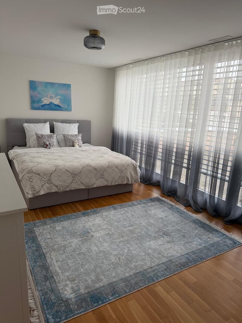 Modern bedroom with a large bed, decorative pillows, a blue and white rug, and a painting on the wall. The room features wooden flooring and large windows with sheer curtains.