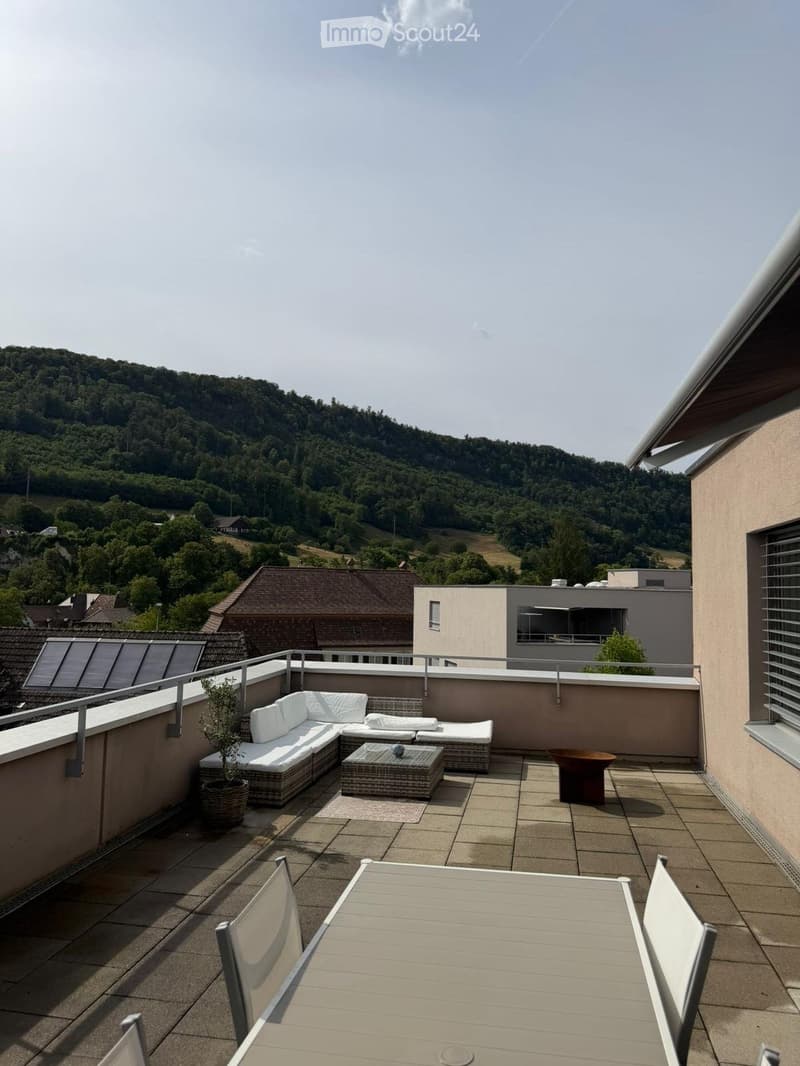 Balcony with a white sectional sofa, coffee table, dining table with chairs, and a potted plant, overlooking a green, hilly landscape.