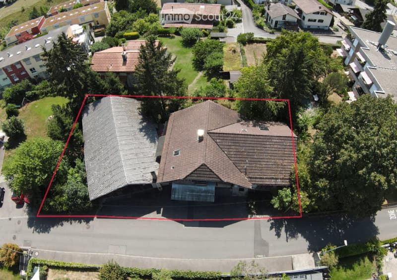 Aerial view of a two-story house with a gray roof and multiple chimneys, surrounded by trees and neighboring houses.