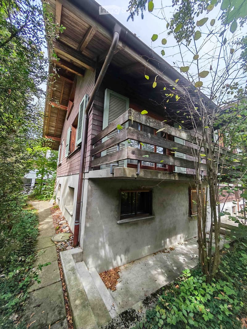 Two-story house with wooden siding and a balcony, surrounded by greenery and trees.