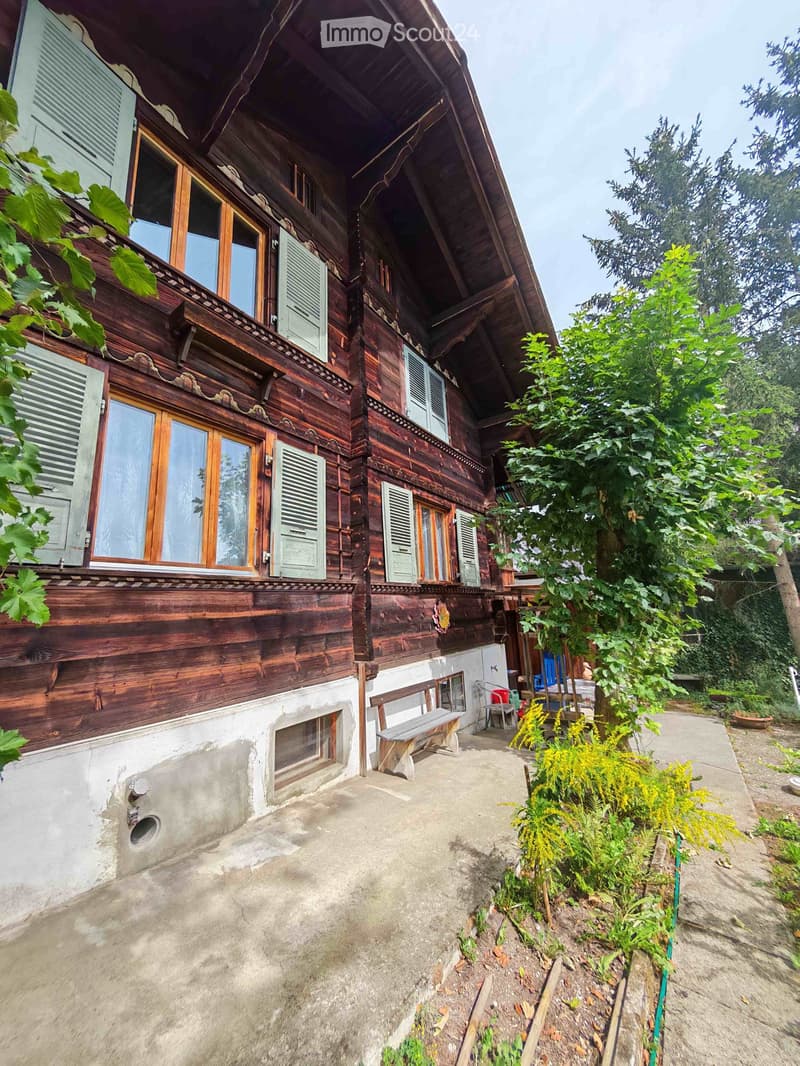 A two-story wooden house with green shutters, a balcony, and a small garden area with a bench and plants.