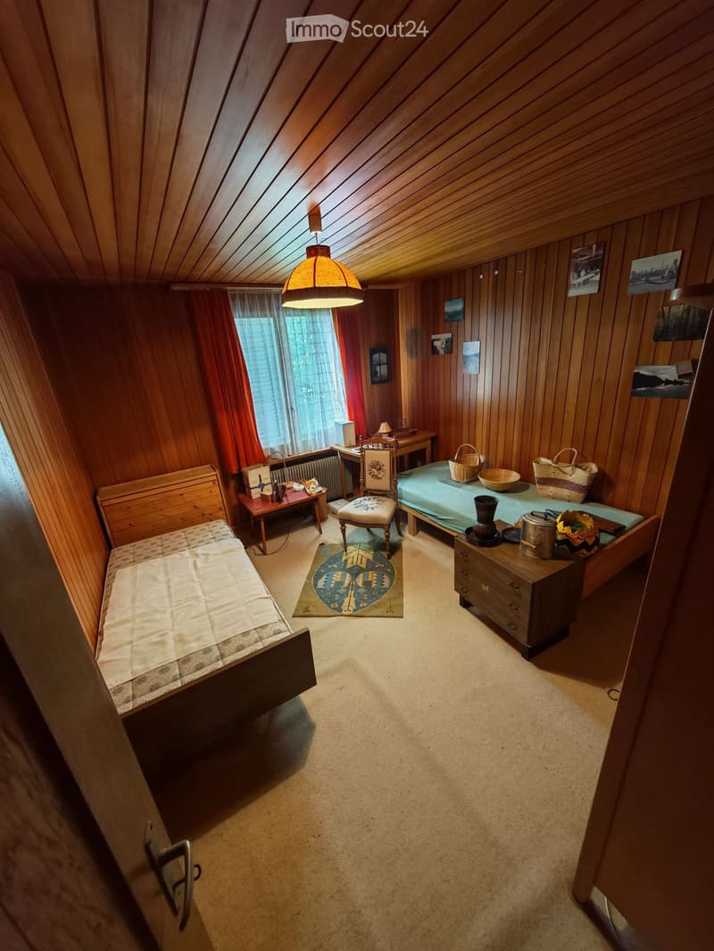 Cozy attic bedroom with wooden walls and slanted ceilings, featuring two single beds, a small table with a lamp, a stool, and decorative baskets.