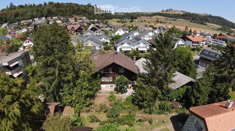 Aerial view of a residential area with multiple houses, surrounded by trees and greenery, with a hilly landscape in the background.