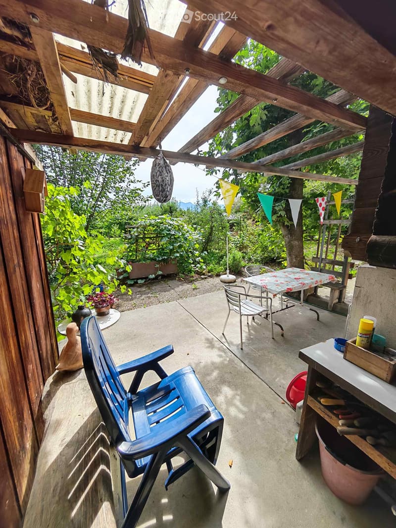 A wooden-beamed balcony with a blue plastic chair, small table with floral tablecloth, and a wooden shelf with cleaning supplies.