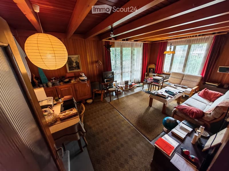 A cozy living room with wooden walls and ceiling, featuring a sofa, coffee table, chairs, books, a globe, and a television. The room has large windows with red curtains and a warm, inviting atmosphere.