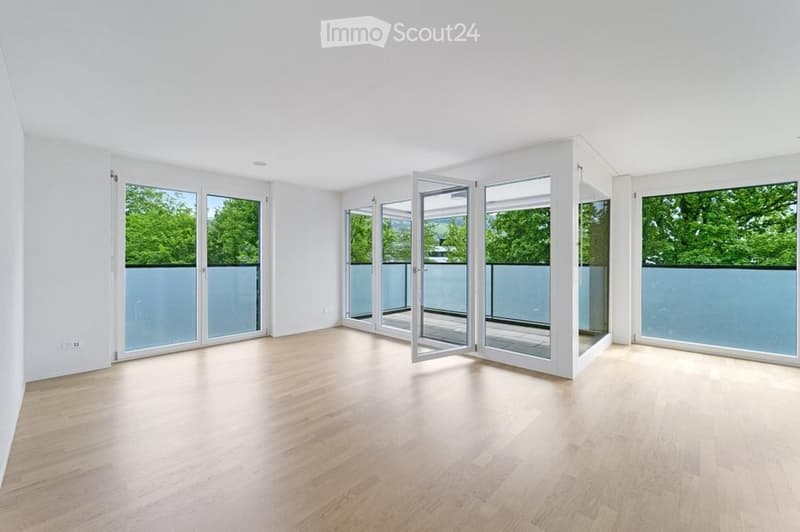 Empty room with light wooden flooring, white walls, and large glass sliding doors leading to a balcony with a view of greenery.