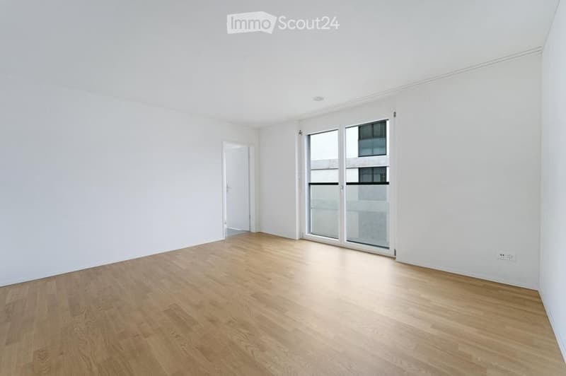 Empty room with white walls, wooden flooring, and large windows with a view of the building exterior.