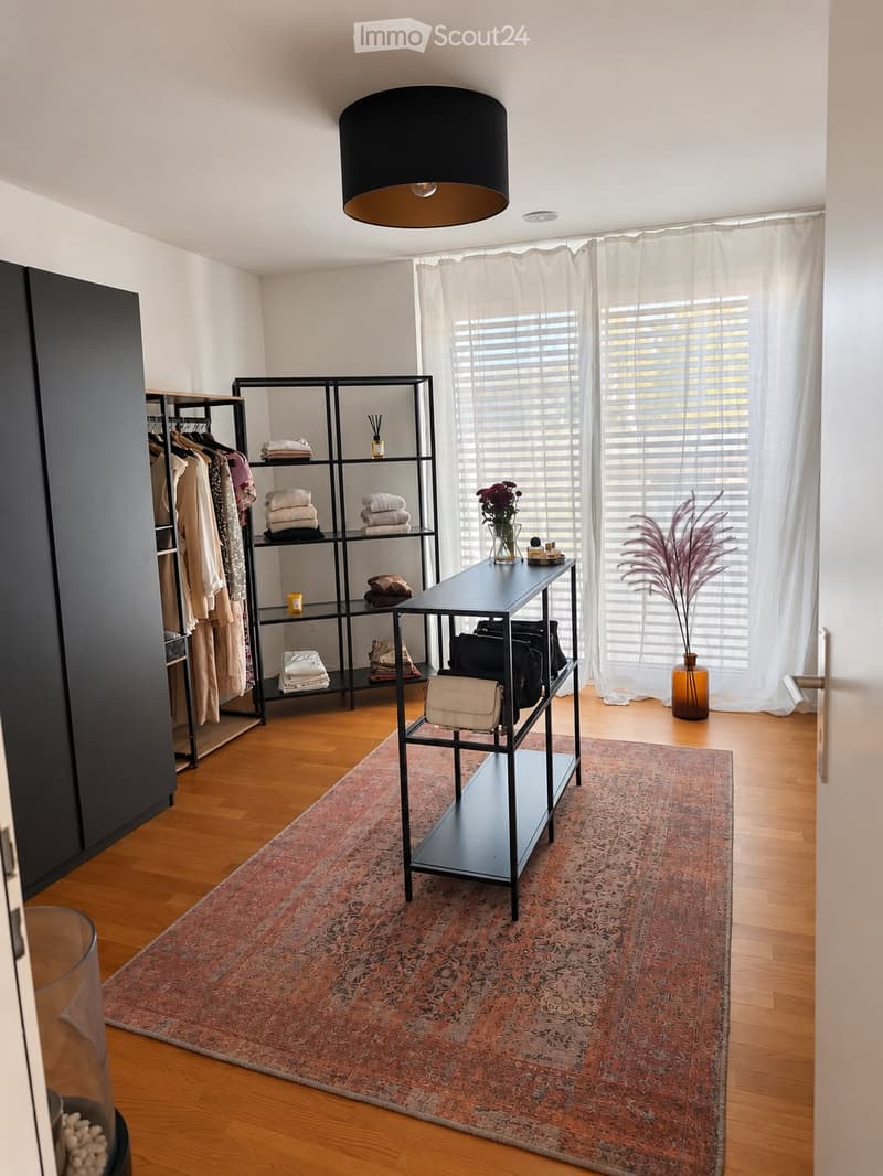 A modern, well-lit room with wooden flooring, a large area rug, a black shelf unit with folded towels and folded clothes, a small black table with a vase of flowers and a decorative item, and a large black pendant lamp hanging from the ceiling.