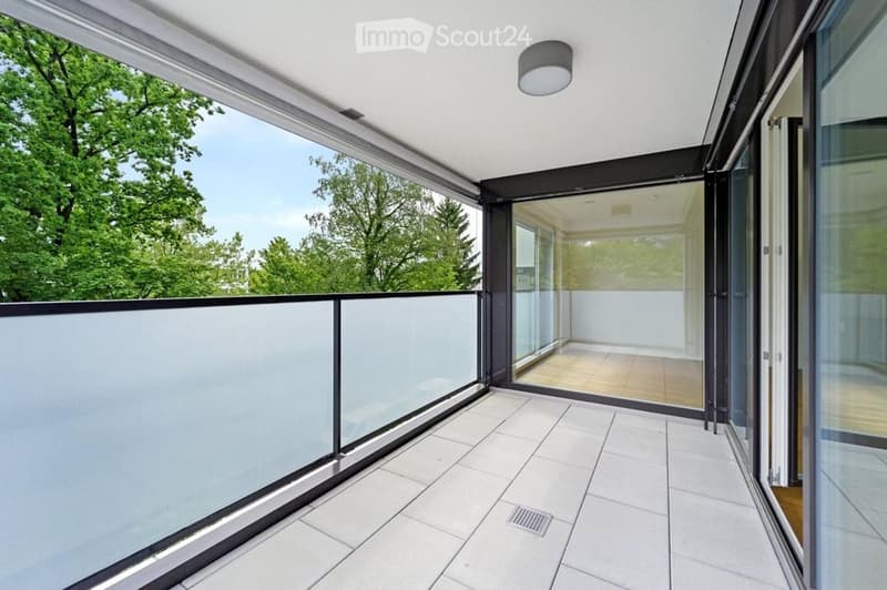 Modern balcony with white tiled flooring, glass railing, and sliding glass doors leading to an interior space.