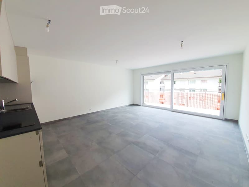 Empty room with grey tiled floor, white walls, and a sliding glass door leading to a balcony. A small kitchen area with a sink and cabinets is visible on the left side.