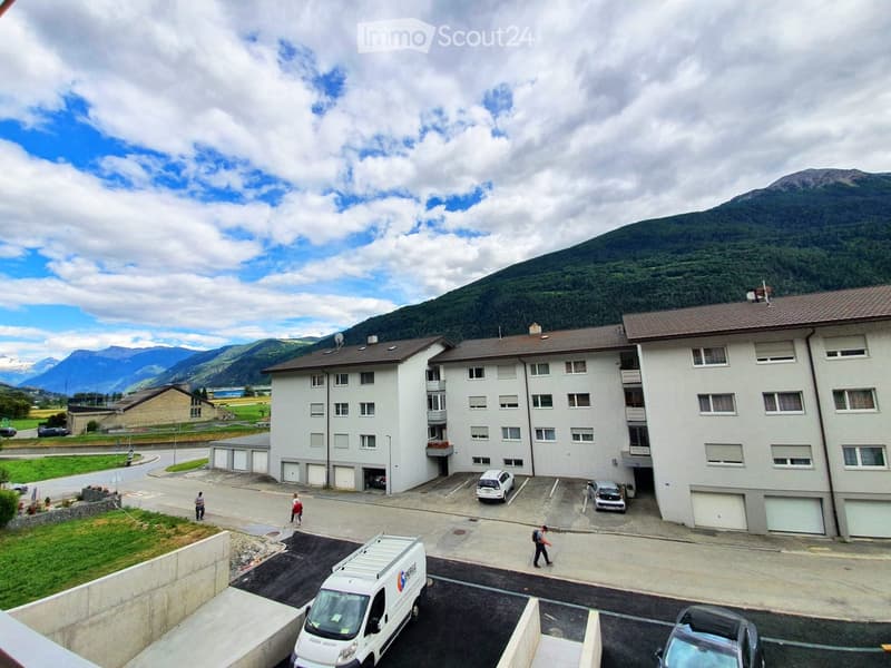Multi-story residential building with multiple apartments, balconies, and parking area, surrounded by a parking lot and mountain backdrop.