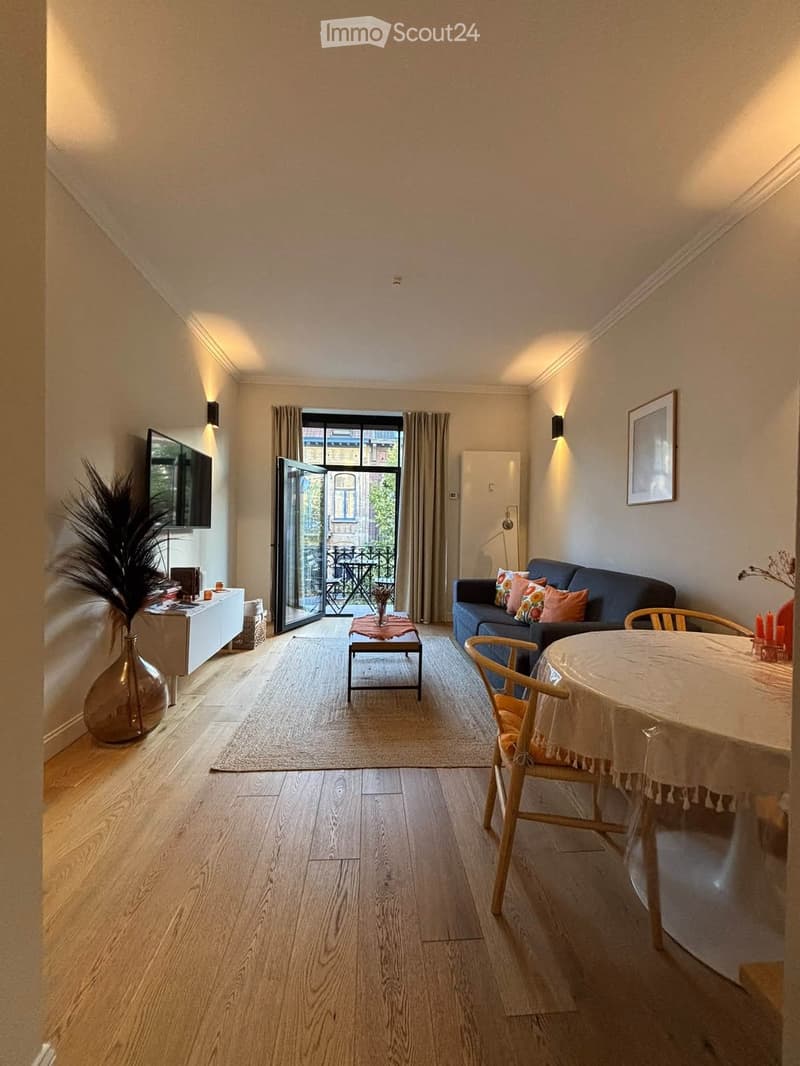 Bright living room with wooden flooring, a sofa, coffee table, dining table, and chairs, with a large window leading to a balcony.