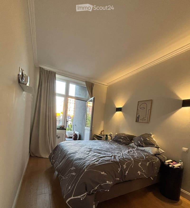 Modern bedroom with a large bed, nightstand, wall-mounted shelf, and a view through an open window to a balcony area.