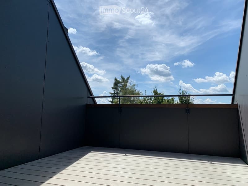 Modern balcony with wooden decking, metal railing, and grey walls, offering a view of trees and a partly cloudy sky.