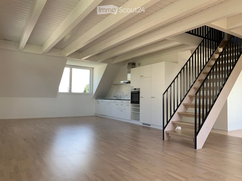 Open-plan space with wooden flooring, white kitchen cabinets, a staircase with wooden steps and black railing, and large windows allowing natural light.