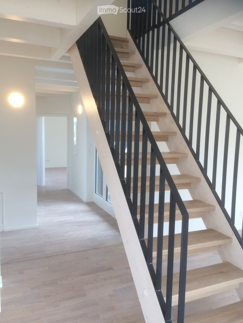 Modern staircase with wooden steps and black metal railing, leading to an open area with light-colored walls and wooden flooring.