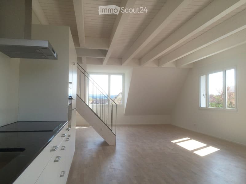 Open-plan space with a modern kitchen area on the left, a staircase leading upwards, and large windows allowing natural light to flood the room. The floor is light-colored wood, and the walls are white.
