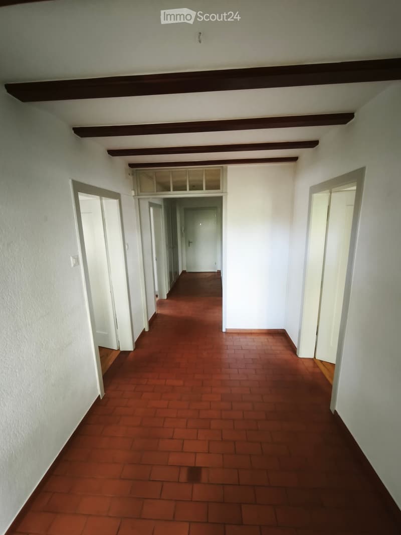 A long, narrow hallway with red brick flooring, white walls, and several open doorways leading to other rooms. The ceiling features exposed wooden beams.