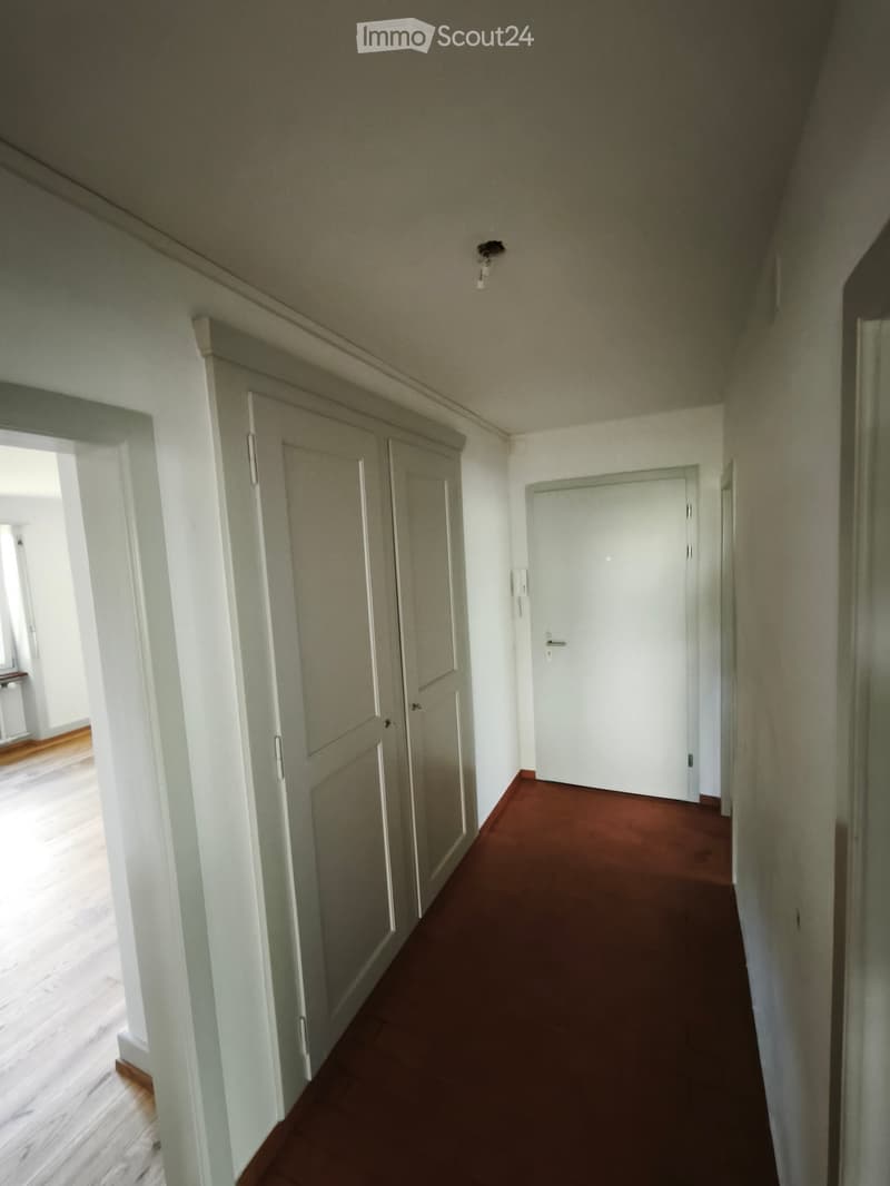 A narrow hallway with white walls, wooden doors, and a red carpeted floor. The hallway features a light fixture on the ceiling and a visible room entrance on the left side.