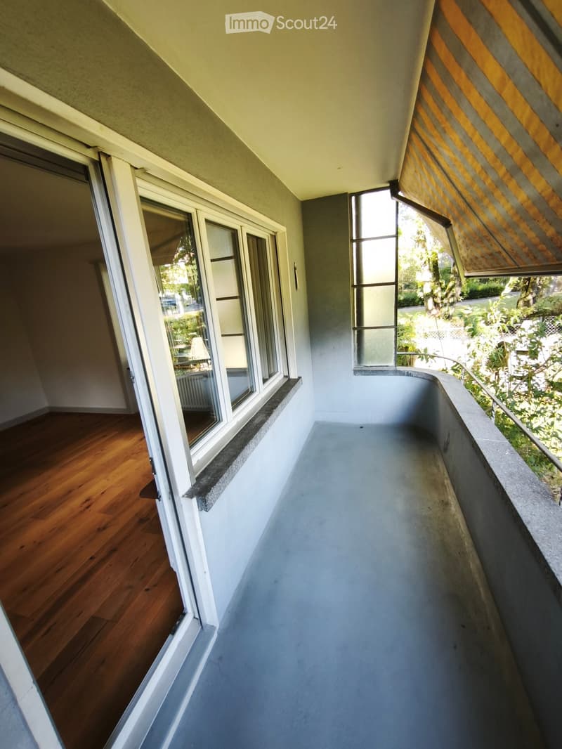 Balcony with wooden floor, large windows, and a view of greenery outside.