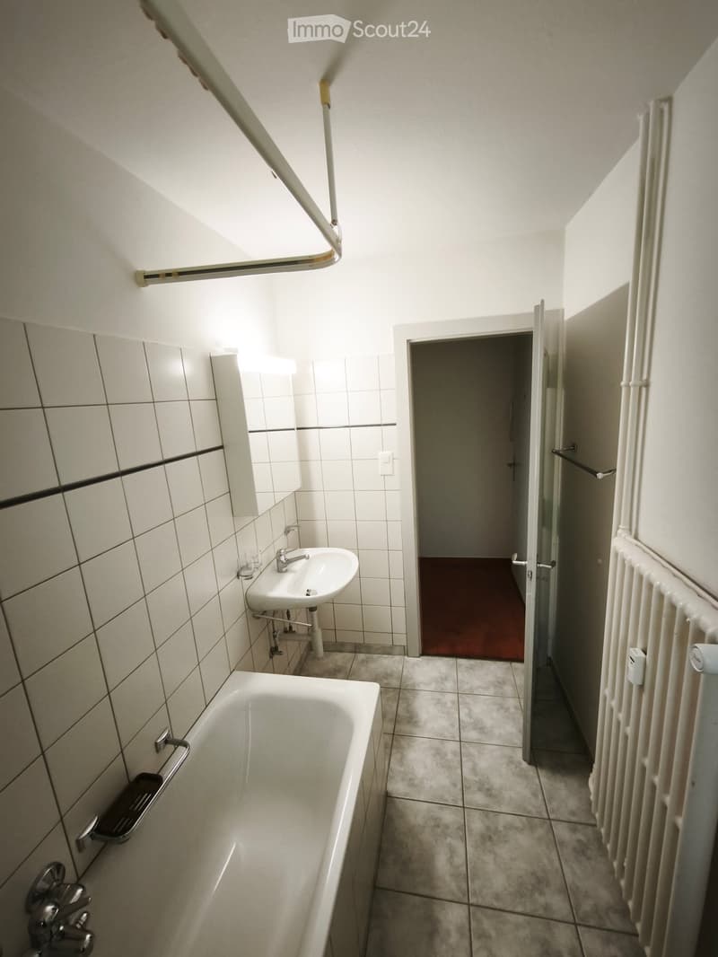 Bathroom with a bathtub, sink, mirror, and radiator. The room has tiled walls and a tiled floor, with an open doorway leading to another room.