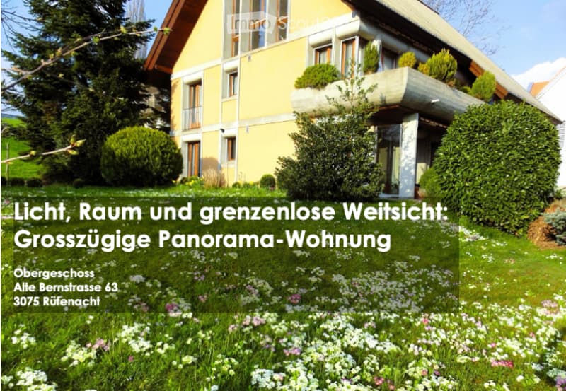 A two-story yellow house with white trim, multiple windows, and a balcony on the upper floor. The house is surrounded by a well-maintained garden with green bushes and flowering plants.