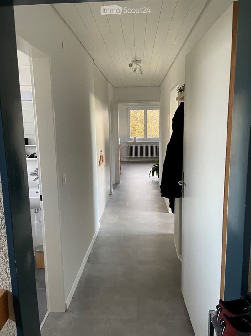A clean, well-lit hallway with white walls, a wooden ceiling, and tiled flooring. A coat is hanging on the right side, and a small plant is visible in the background near a window.