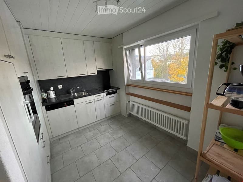 Modern kitchen with white cabinets, grey countertops, tiled floor, and a window overlooking the outside. Includes a sink, dishwasher, oven, refrigerator, and a small wooden shelf with a bowl and toaster.