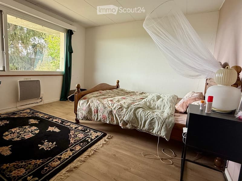 Cozy bedroom with wooden floor, large window with green curtain, bed with floral duvet, nightstand with lamp, radiator, and decorative rug.