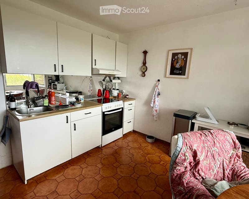 A small kitchen with white cabinets, a sink, an oven, microwave, and a small table with a chair. The floor is tiled in a hexagonal pattern, and there is a picture frame and a clock on the wall.
