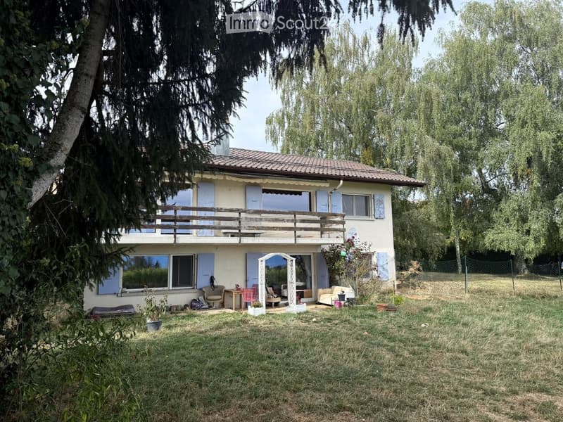 Two-story house with a tiled roof, light-colored exterior, multiple windows, and a wooden balcony on the upper level. The surrounding area includes a grassy yard with trees and outdoor furniture.