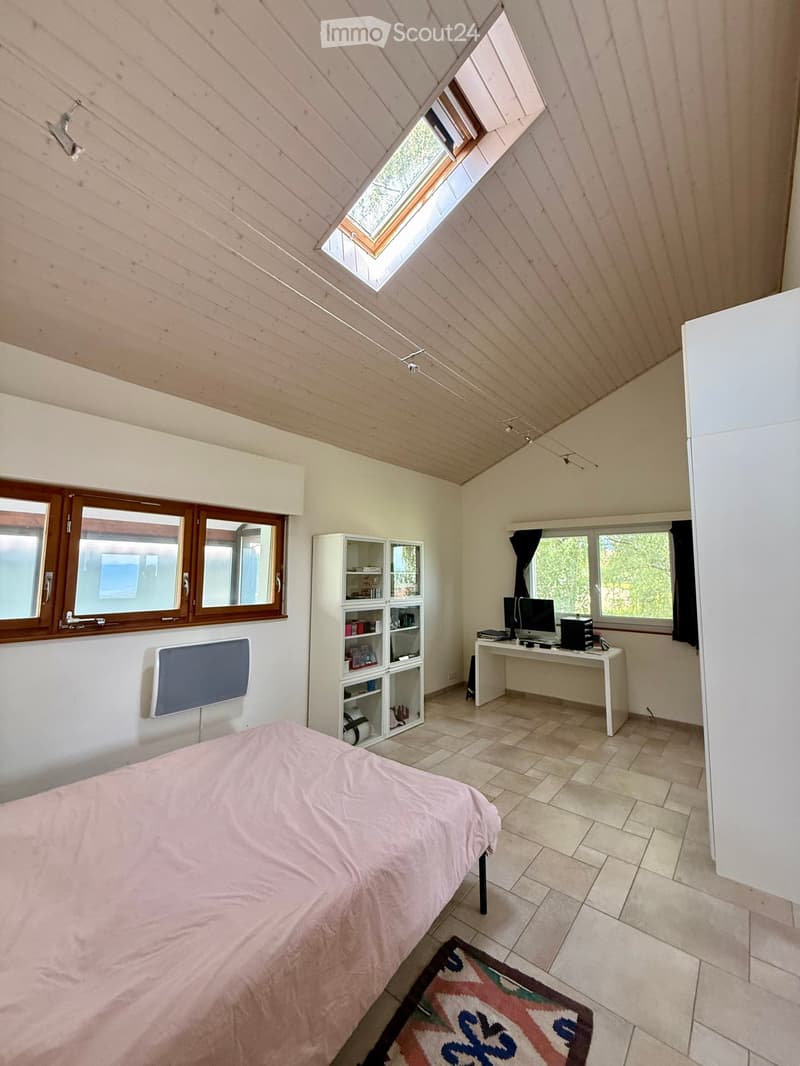 Bright bedroom with a bed, desk, computer, shelf, and mirror. The room features wooden ceilings and tiled flooring.