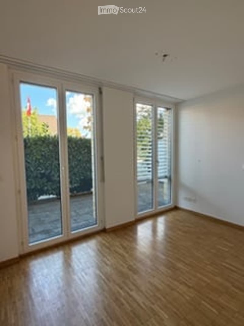 Empty room with wooden flooring, large glass sliding doors leading to a balcony, white walls, and bright natural light.