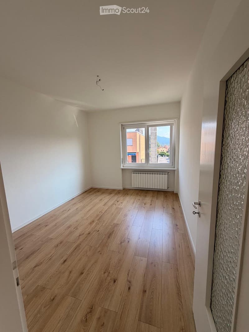 Empty room with wooden flooring, white walls, and a window with a view of a building and mountains.