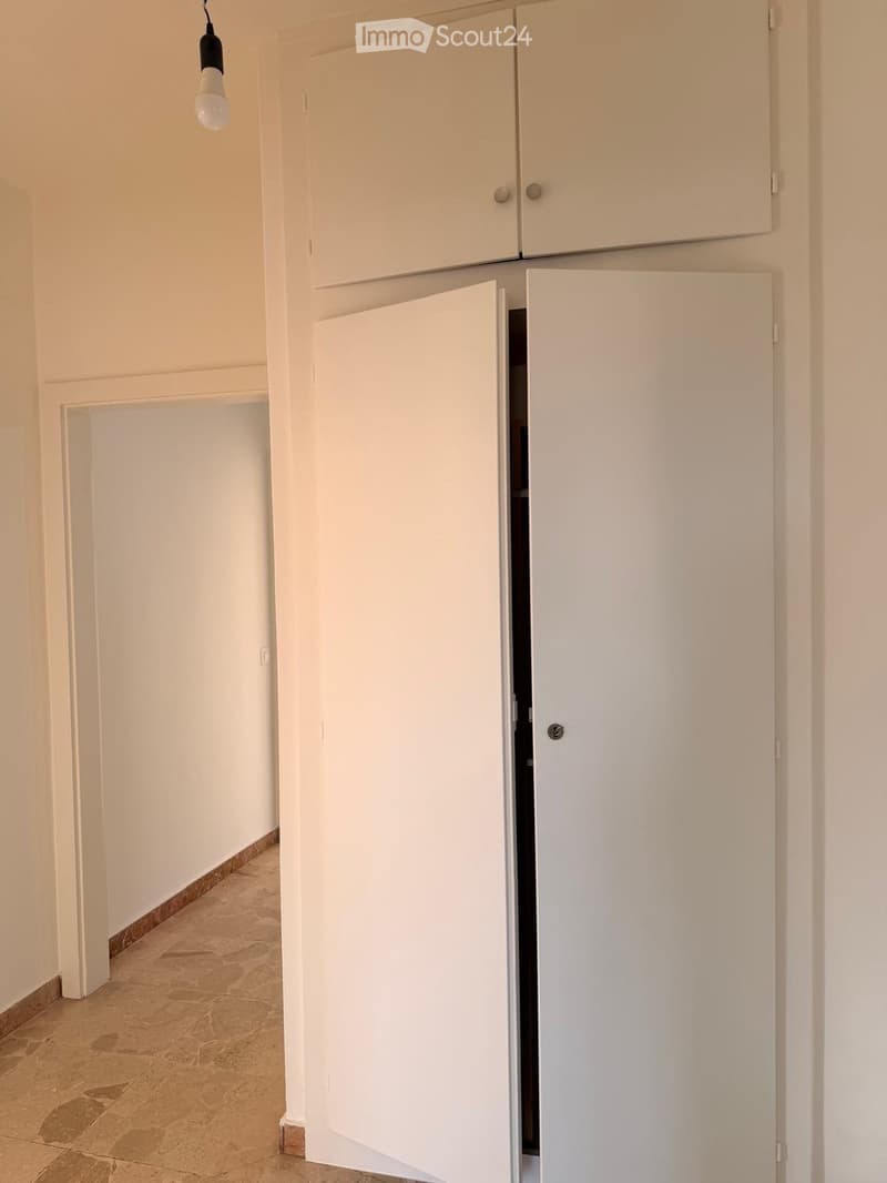 A narrow hallway with beige walls, tiled floor, and white cabinetry on one side. A light fixture hangs from the ceiling, and a doorway leads to another room.