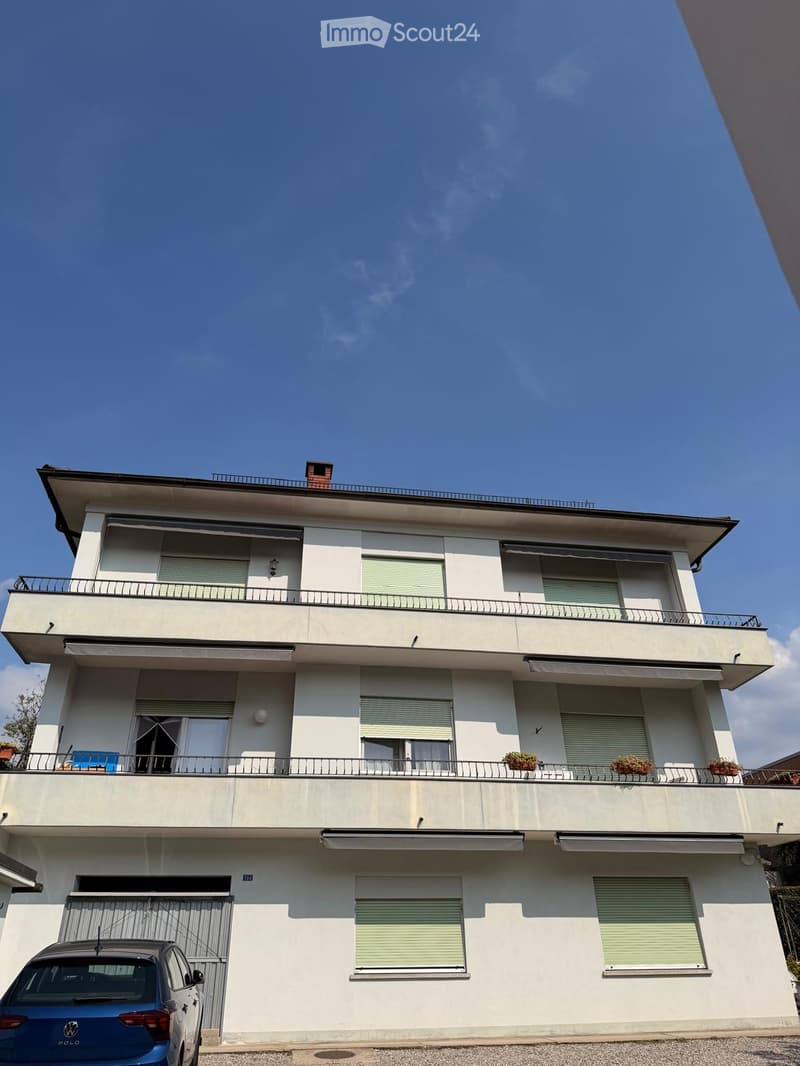 Three-story house with white exterior, multiple balconies, and a rooftop terrace. Features include closed shutters, potted plants on balconies, and a parked car in the driveway.