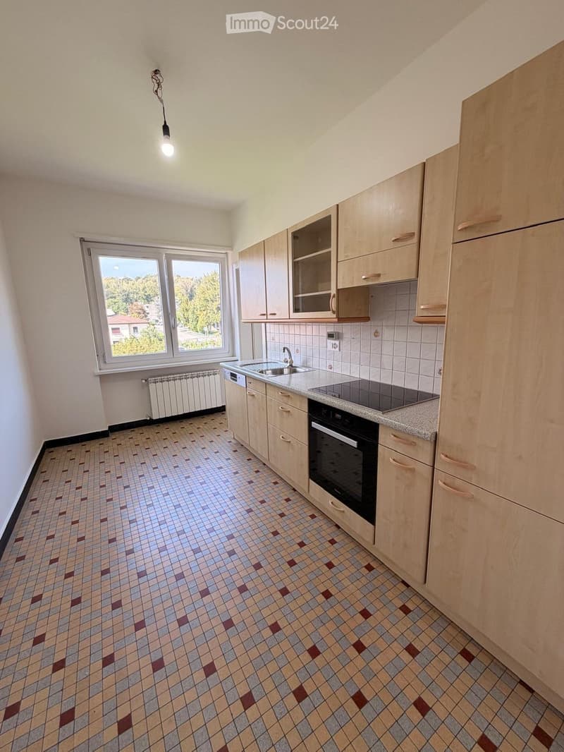 Empty kitchen with light wooden cabinets, tiled floor, and a window overlooking a green area.