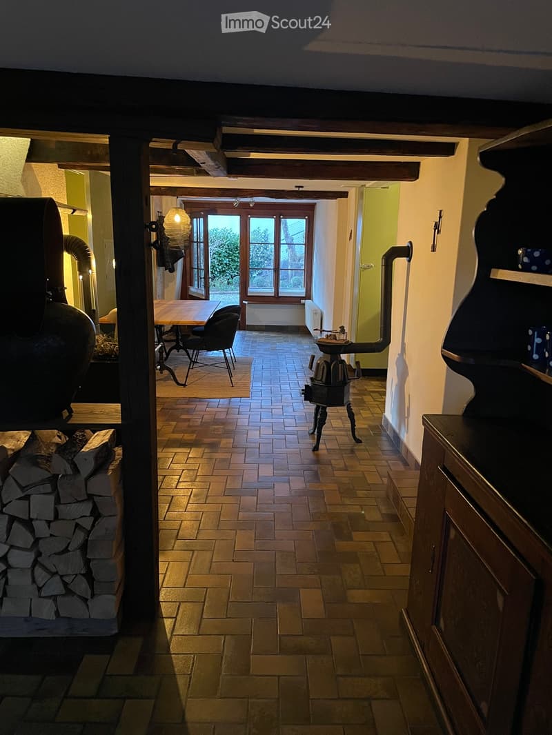 A well-lit hallway with wooden beams, tiled floor, and a view into a dining area with a table and chairs. Features a wood stove and a stack of firewood on the left side.
